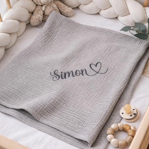May include: A light gray muslin baby blanket featuring the embroidered name Simon in a dark gray cursive script followed by a small heart outline. The blanket is folded and placed inside a wooden crib with a white braided bumper and a teddy bear.