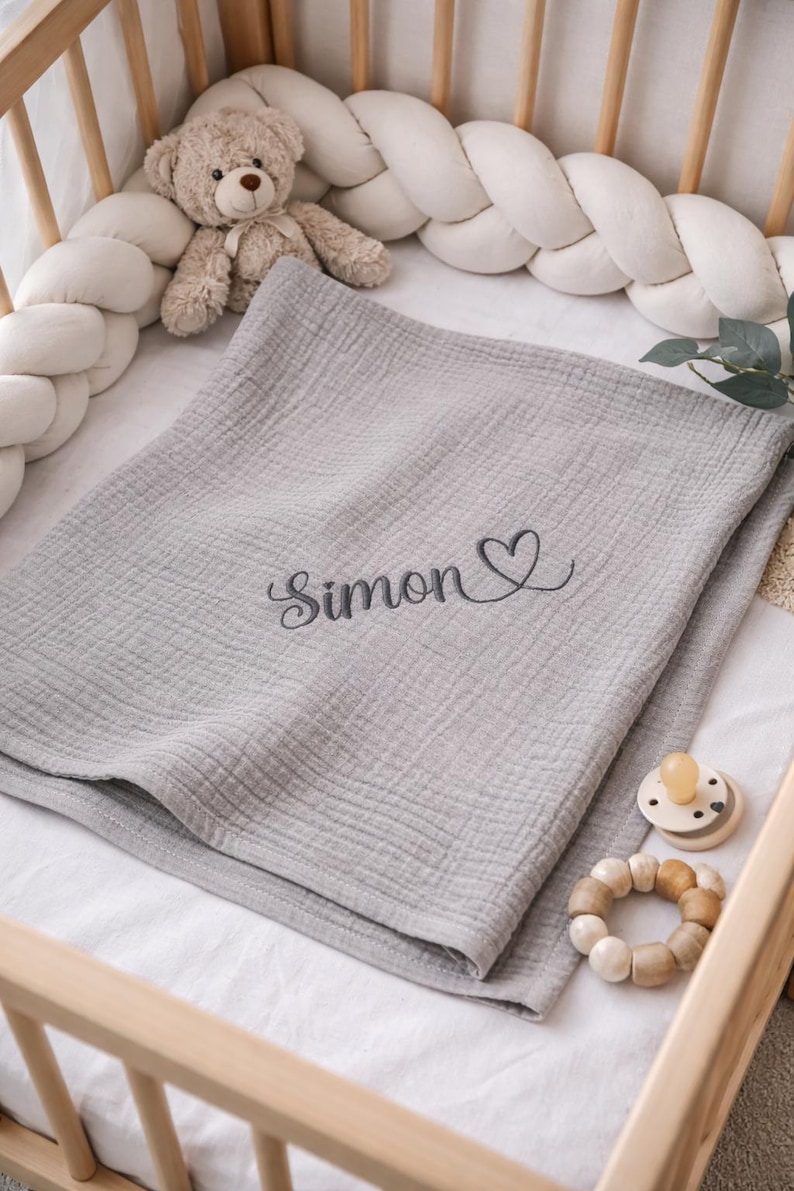 May include: A light gray muslin baby blanket featuring the embroidered name Simon in a dark gray cursive script followed by a small heart outline. The blanket is folded and placed inside a wooden crib with a white braided bumper and a teddy bear.