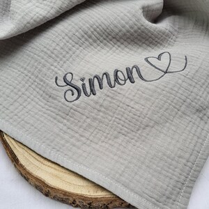 May include: A light gray muslin cloth featuring a textured, crinkled fabric surface. The name Simon is embroidered in dark gray cursive script, followed by a small heart outline. A beige pacifier is visible near the edge of the fabric.