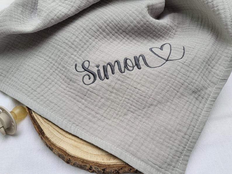 May include: A light gray muslin cloth featuring a textured, crinkled fabric surface. The name Simon is embroidered in dark gray cursive script, followed by a small heart outline. A beige pacifier is visible near the edge of the fabric.