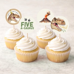 Puede incluir: Cuatro cupcakes con glaseado blanco y adornos con temática de dinosaurios. Los adornos presentan un dinosaurio con gorro de fiesta, un volcán y el texto "FIVE A-SAURUS" y "ROAR!" en verde y marrón. Los cupcakes están en moldes de papel amarillos.