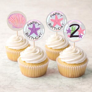 Op de afbeelding: Vier cupcakes met witte frosting en decoratieve toppers. De toppers bevatten een roze zeeschelp, een paarse zeester, een roze zeester en een topper met het nummer 2. De toppers hebben de tekst "Happy Birthday".