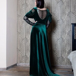 May include: A long, emerald green velvet dress with a sheer, sparkly lace back panel and long sleeves. The dress has a fitted bodice and a flowing skirt.