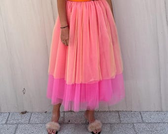 May include: A bright orange short-sleeved top with a twisted front detail paired with a layered midi-length tulle skirt featuring a soft peach upper section and a vibrant hot pink bottom tier, worn with light-colored open-toe slide sandals.