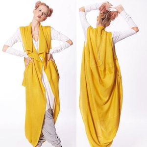 Può includere: Un lungo gilet giallo senza maniche con un design drappeggiato. Il gilet è indossato sopra una camicia bianca e pantaloni grigi.