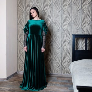 May include: A woman wearing a long, emerald green velvet dress with lace sleeves. The dress has a fitted bodice and a flowing skirt. She is standing in a room with a bed in the background.