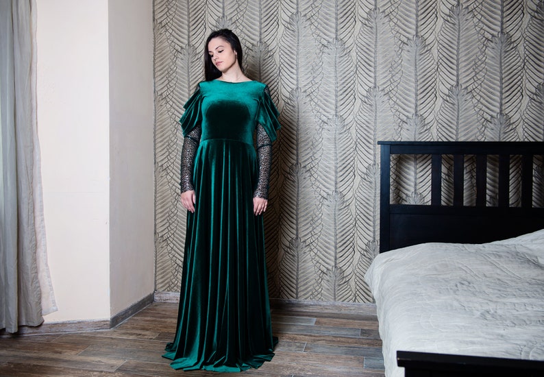 May include: A woman wearing a long, emerald green velvet dress with lace sleeves. The dress has a fitted bodice and a flowing skirt. She is standing in a room with a bed in the background.