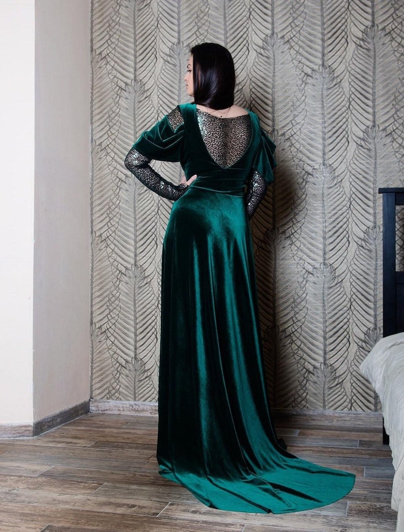 May include: A long, emerald green velvet dress with a sheer, sparkly lace back panel and long sleeves. The dress has a fitted bodice and a flowing skirt.