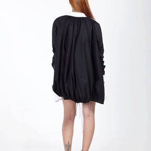 K&ouml;nnte beinhalten: Eine schwarze Oversize-Jacke mit gerafftem Saum und wei&szlig;em Kragen, getragen &uuml;ber einem wei&szlig;en Hemd. Die Jacke hat lange &Auml;rmel mit wei&szlig;en Manschetten und eine lockere, volumin&ouml;se Silhouette, die den R&uuml;cken bedeckt, kombiniert mit schwarzen Stiefeletten.