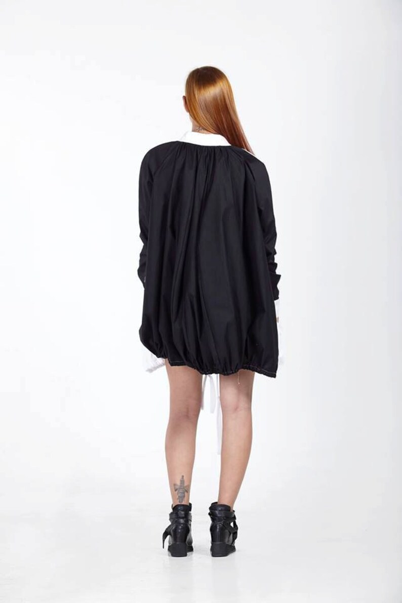 K&ouml;nnte beinhalten: Eine schwarze Oversize-Jacke mit gerafftem Saum und wei&szlig;em Kragen, getragen &uuml;ber einem wei&szlig;en Hemd. Die Jacke hat lange &Auml;rmel mit wei&szlig;en Manschetten und eine lockere, volumin&ouml;se Silhouette, die den R&uuml;cken bedeckt, kombiniert mit schwarzen Stiefeletten.