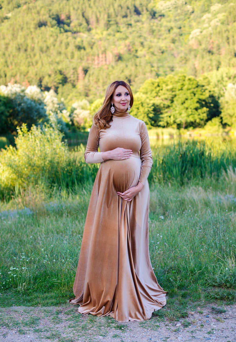 May include: A floor-length, long-sleeved velvet dress in a light tan color featuring a high turtleneck collar and a fitted bodice that transitions into a full, flowing skirt. The fabric has a soft sheen, and the dress is worn by a person.