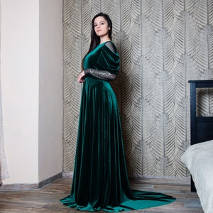 May include: A woman wearing a long, emerald green velvet dress with a fitted bodice and a flowing skirt. The dress has long sleeves with a sparkly detail on the shoulders. She is standing in a room with a bed in the background.