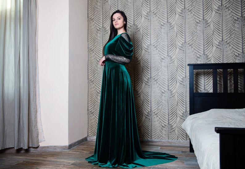 May include: A woman wearing a long, emerald green velvet dress with a fitted bodice and a flowing skirt. The dress has long sleeves with a sparkly detail on the shoulders. She is standing in a room with a bed in the background.