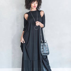 May include: A floor-length black dress with cold-shoulder cutouts, long sleeves, and a sheer bottom panel featuring subtle vertical stud detailing. The garment is paired with a long silver chain necklace and a black handbag with silver grommets.