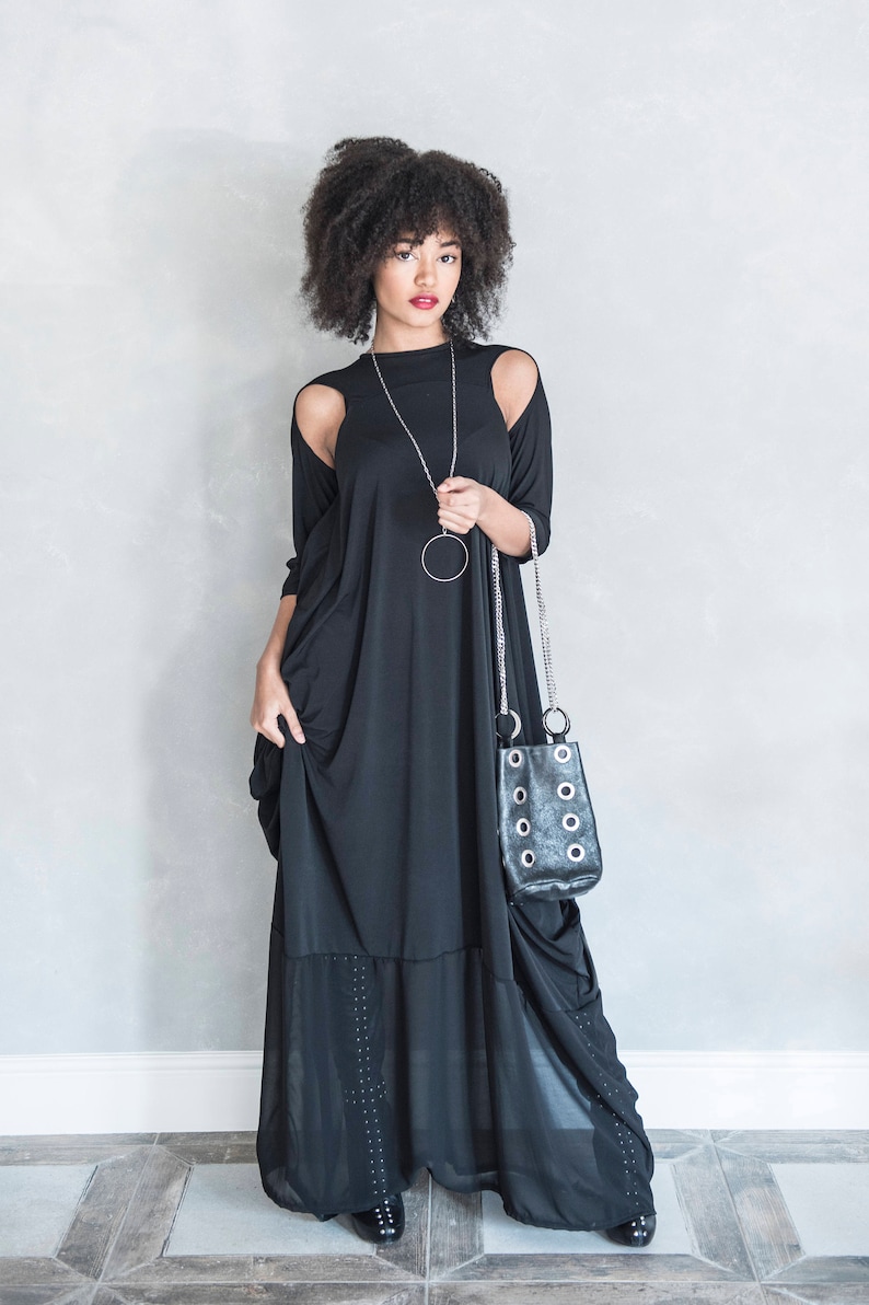 May include: A floor-length black dress with cold-shoulder cutouts, long sleeves, and a sheer bottom panel featuring subtle vertical stud detailing. The garment is paired with a long silver chain necklace and a black handbag with silver grommets.