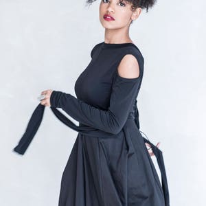 Pu&ograve; includere: Un abito nero a maniche lunghe con aperture sulle spalle, scollo rotondo e lunghi nastri di tessuto che partono dalla vita. Il capo presenta un corpetto aderente che si trasforma in una gonna ampia e fluida, creando una silhouette distinta.