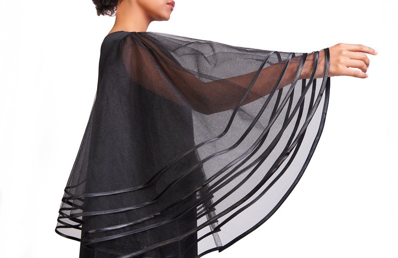 May include: A black sheer capelet featuring wide, flowing sleeves made of translucent mesh fabric. The hem of the capelet and the edges of the sleeves are finished with multiple horizontal bands of solid black ribbon trim, creating a layered effect.