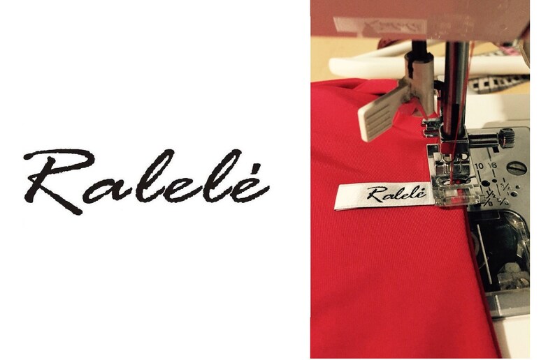 May include: A white fabric clothing label with the black text Ralel&eacute; printed in a cursive script font. The label is being sewn onto a piece of bright red fabric using a silver metal sewing machine presser foot and needle mechanism.