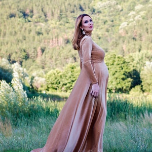 May include: A floor-length, long-sleeved velvet dress in a warm tan color. The garment features a high mock neck, a fitted bodice that accommodates a pregnancy bump, and a flowing, full-length skirt that pools slightly at the bottom hemline.