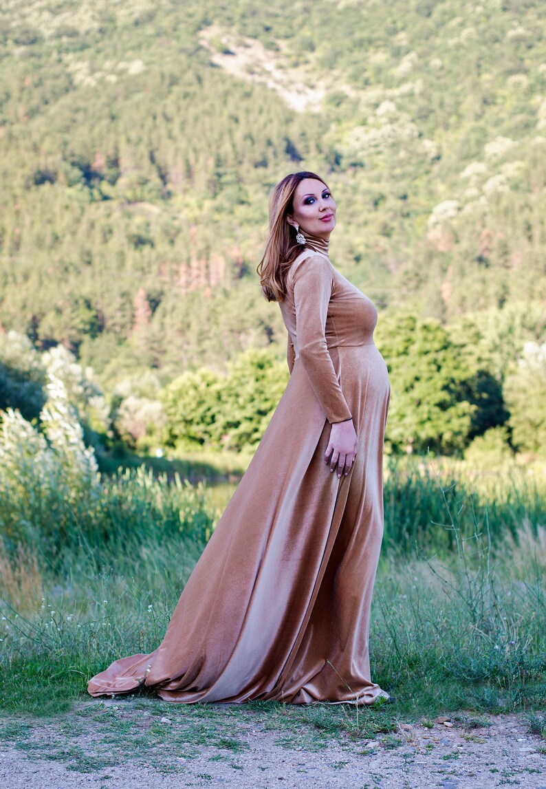 May include: A floor-length, long-sleeved velvet dress in a warm tan color. The garment features a high mock neck, a fitted bodice that accommodates a pregnancy bump, and a flowing, full-length skirt that pools slightly at the bottom hemline.