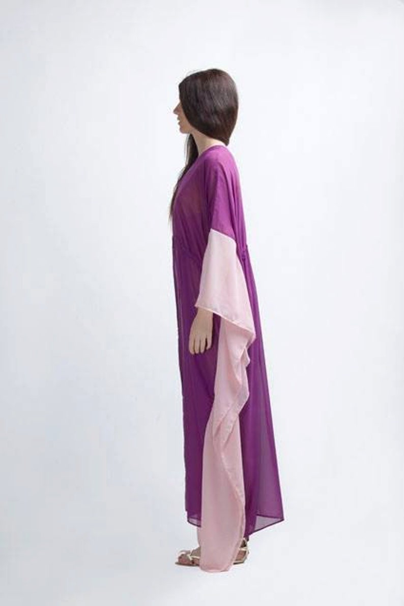 Puede incluir: Un vestido largo y fluido de color p&uacute;rpura con mangas anchas en un rosa claro contrastante. El vestido est&aacute; hecho de una tela transparente y tiene un ajuste holgado y c&oacute;modo. La persona lleva sandalias.
