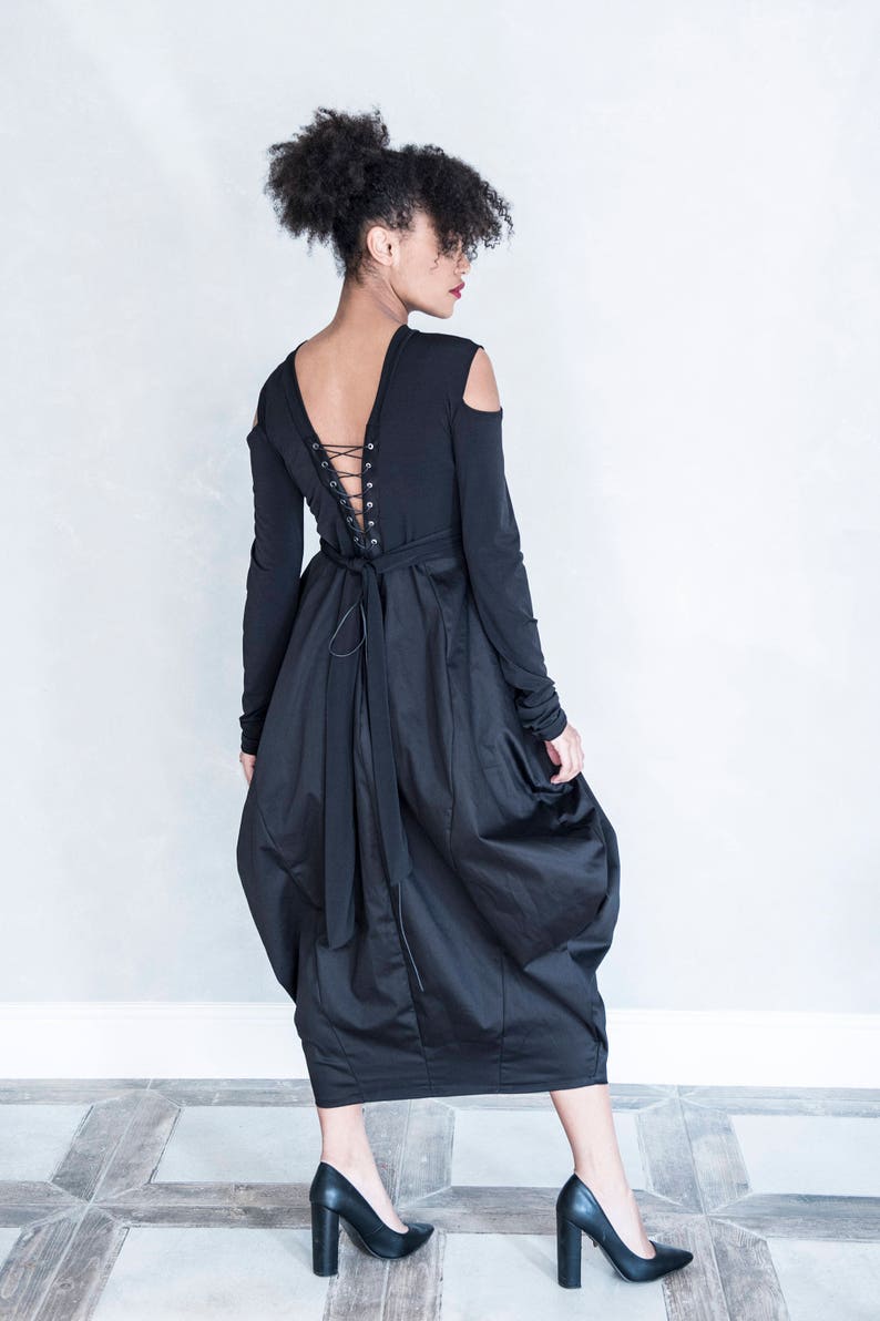 Pu&ograve; includere: Un abito nero a maniche lunghe con aperture sulle spalle, scollo a V profondo con chiusura a lacci stile corsetto sulla schiena e una gonna voluminosa e arricciata di lunghezza midi, abbinato a d&eacute;collet&eacute; nere con tacco alto.