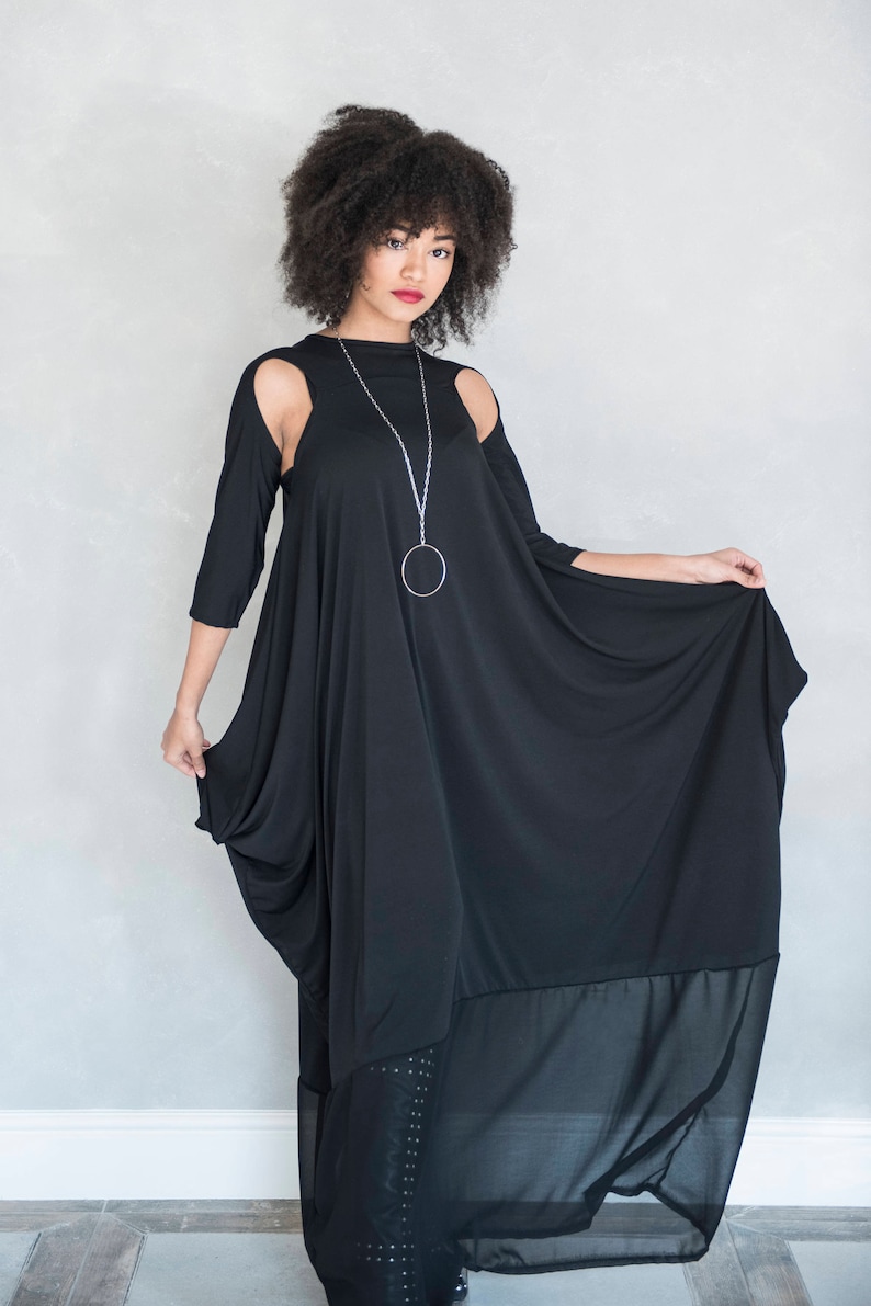 May include: A black floor-length dress featuring three-quarter length sleeves with shoulder cutouts, a loose draped silhouette, and a sheer bottom panel. A long silver chain necklace with a large circular pendant is worn with the garment.
