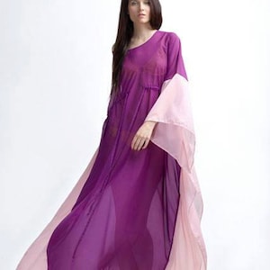 Puede incluir: Un vestido largo y fluido en tonos morados y rosas. El vestido presenta un dise&ntilde;o de un solo hombro con mangas anchas y acampanadas. La tela transparente crea un efecto de capas, con la capa inferior morada m&aacute;s oscura visible a trav&eacute;s de la capa superior rosa m&aacute;s clara.