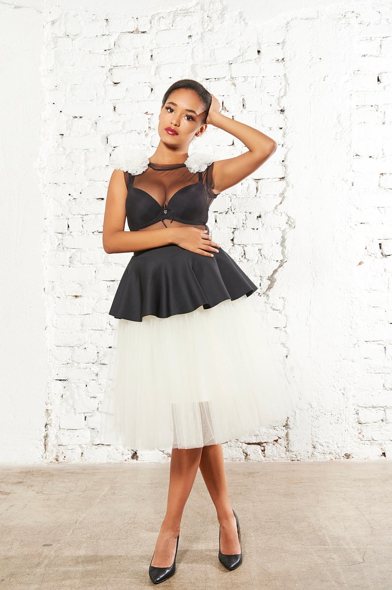May include: A black sleeveless top featuring a sheer mesh neckline, ruffled white shoulder accents, and a structured peplum waist, paired with a knee-length white tulle skirt and black pointed-toe heels, creating a layered ensemble.
