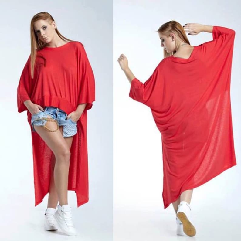 May include: A bright red high-low tunic top featuring a wide, loose-fitting silhouette, short dolman sleeves, and a rounded neckline. The garment is longer in the back than in the front, creating a dramatic draped effect with a soft, flowing fabric.