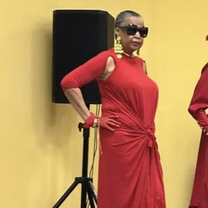 May include: A vibrant red floor-length dress featuring three-quarter length sleeves with shoulder cutouts, a knotted waist detail, and a flowing skirt. The ensemble is paired with matching red gloves, gold dangling earrings, and black sunglasses.