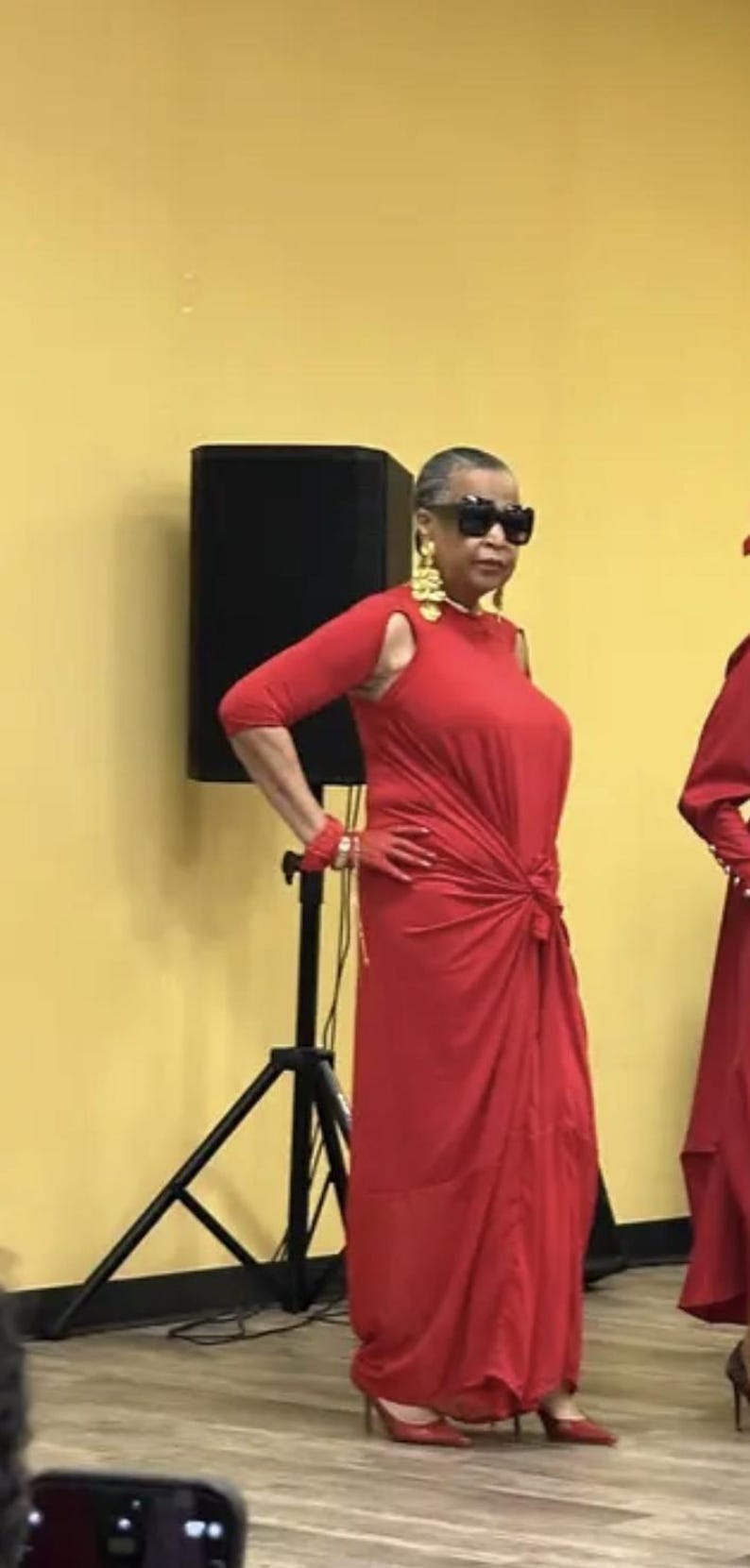 May include: A vibrant red floor-length dress featuring three-quarter length sleeves with shoulder cutouts, a knotted waist detail, and a flowing skirt. The ensemble is paired with matching red gloves, gold dangling earrings, and black sunglasses.