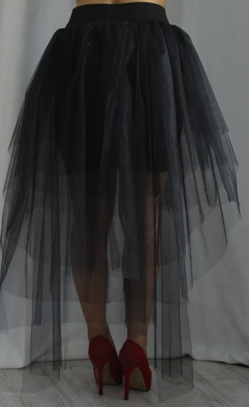 May include: A black tulle skirt featuring a high-low hemline with multiple layers of sheer fabric and a solid black waistband. The skirt is paired with red high-heeled shoes, creating a distinct contrast between the dark skirt and the bright footwear.