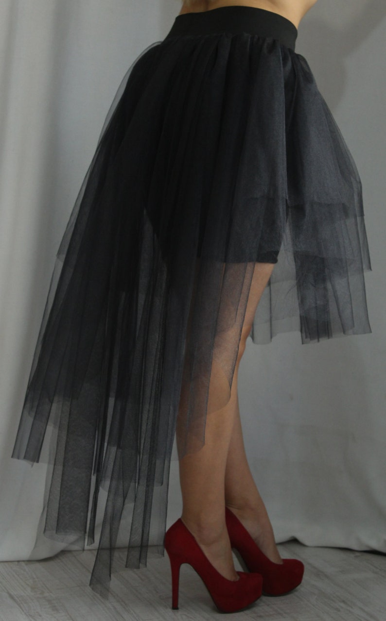 May include: A black high-low skirt made of layered tulle fabric featuring a solid black waistband. The skirt has a shorter front hemline and a longer, flowing back hemline that drapes down, paired with red high-heeled pumps with platform soles.