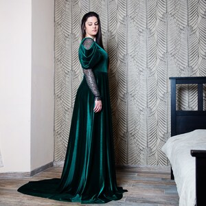 May include: A woman wearing a long, emerald green velvet gown with puffed sleeves and a long train. The gown has a fitted bodice and a flowing skirt. The woman is standing in a room with a patterned wallpaper and a bed in the background.