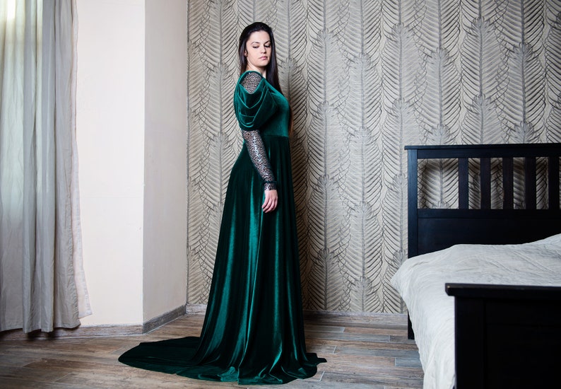 May include: A woman wearing a long, emerald green velvet gown with puffed sleeves and a long train. The gown has a fitted bodice and a flowing skirt. The woman is standing in a room with a patterned wallpaper and a bed in the background.