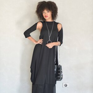May include: A black long-sleeved dress with shoulder cutouts, featuring a layered hemline with a sheer panel. The outfit is accessorized with a long silver chain necklace with a circular pendant and a black handbag with silver grommet details.
