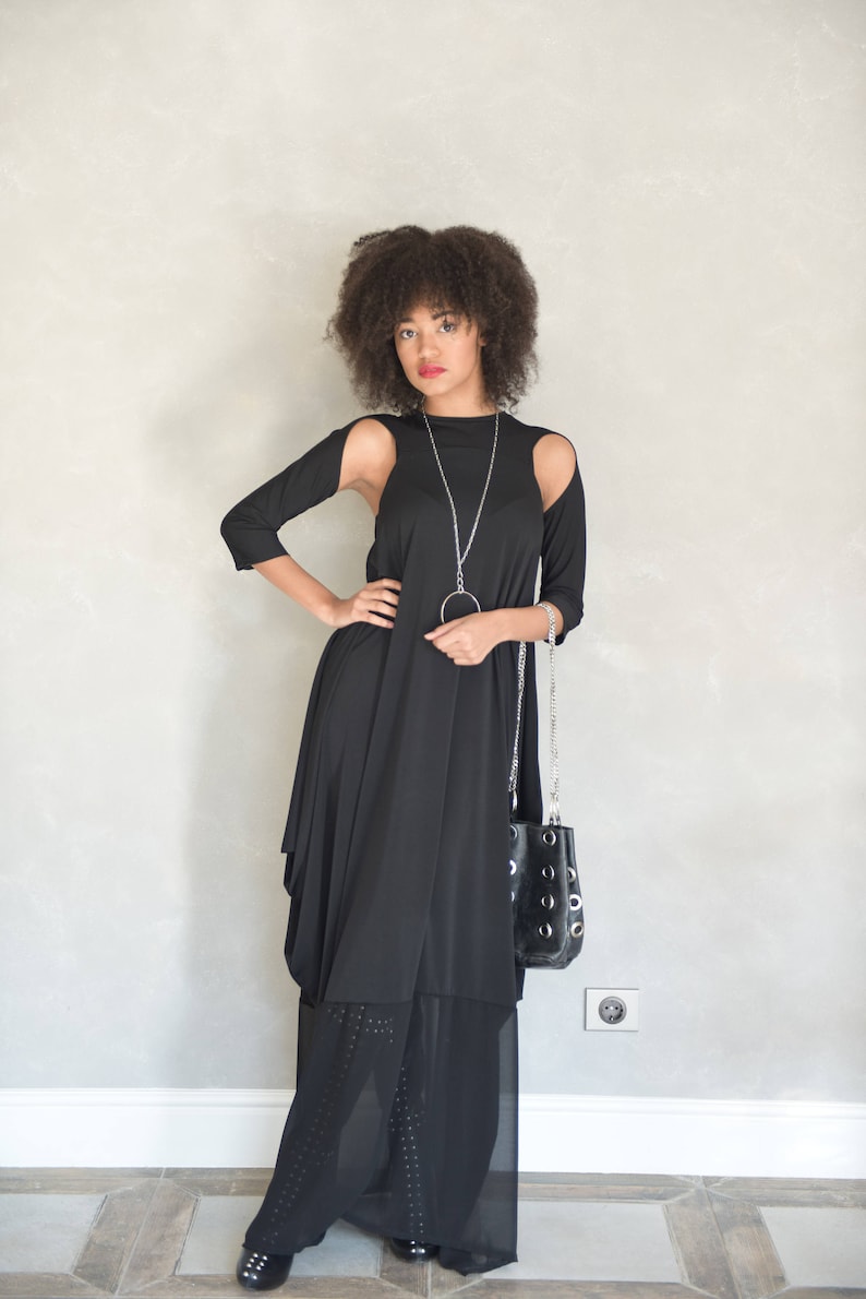 May include: A black long-sleeved dress with shoulder cutouts, featuring a layered hemline with a sheer panel. The outfit is accessorized with a long silver chain necklace with a circular pendant and a black handbag with silver grommet details.