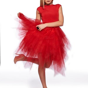 May include: A vibrant red dress featuring a fitted bodice with short sleeves and a voluminous, multi-layered tulle skirt that falls in an asymmetrical, high-low hemline. The garment is paired with neutral-toned pointed-toe high heels.