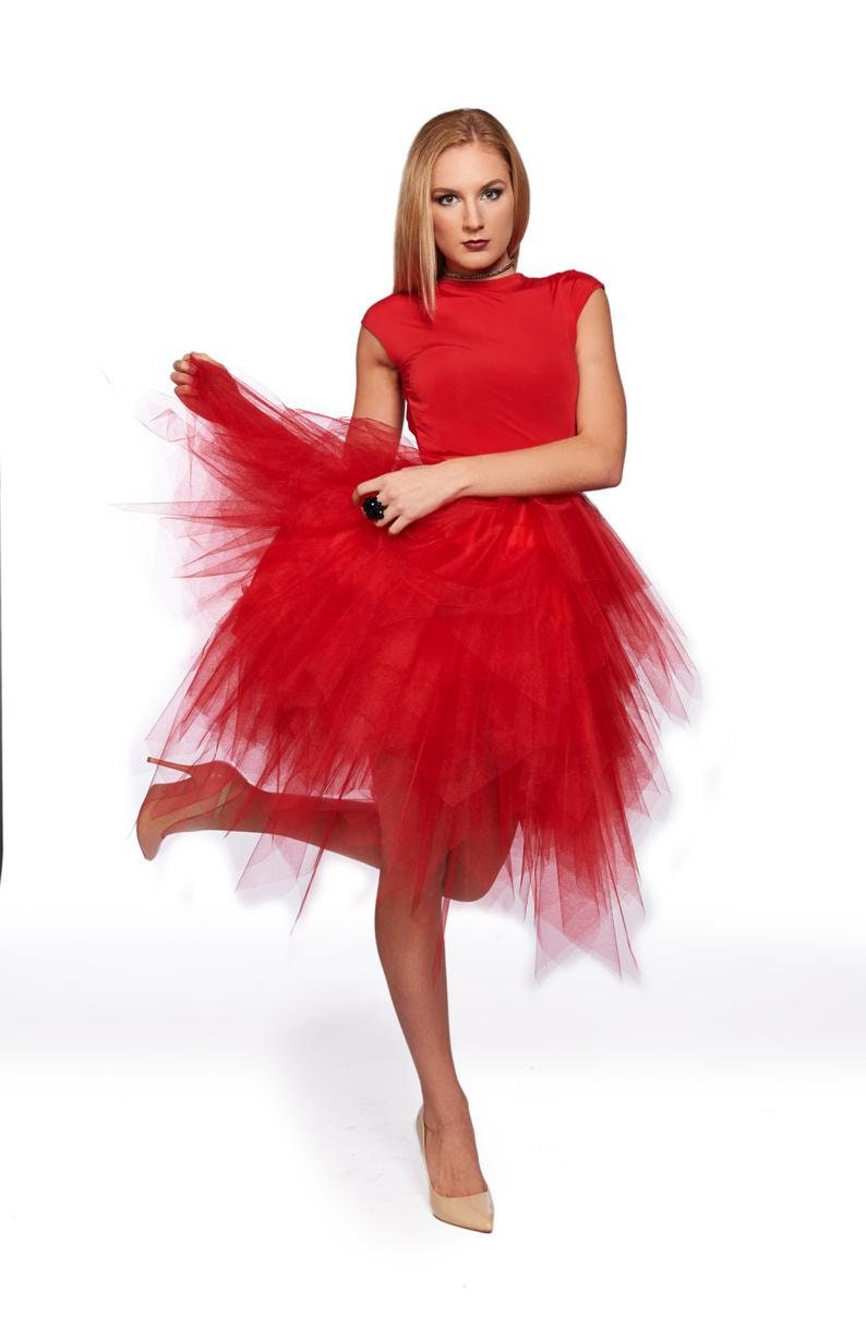 May include: A vibrant red dress featuring a fitted bodice with short sleeves and a voluminous, multi-layered tulle skirt that falls in an asymmetrical, high-low hemline. The garment is paired with neutral-toned pointed-toe high heels.
