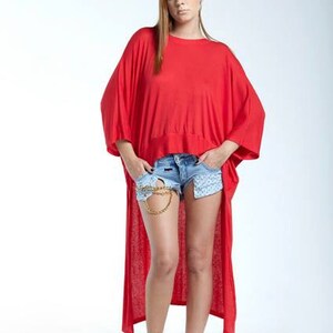 May include: A bright red high-low tunic top with short wide sleeves and a round neckline, paired with light blue denim shorts featuring a gold chain detail at the waist and white high-top sneakers. The tunic has a loose, flowing fit.