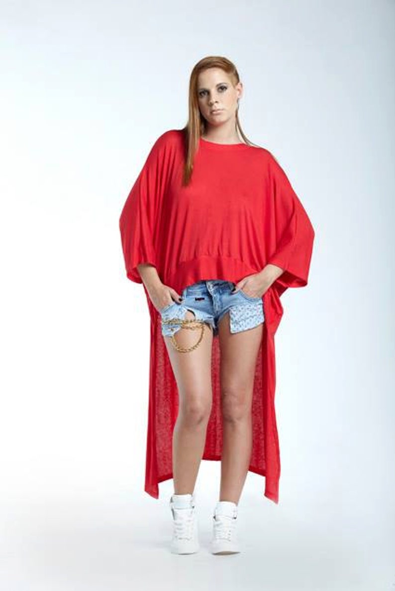 May include: A bright red high-low tunic top with short wide sleeves and a round neckline, paired with light blue denim shorts featuring a gold chain detail at the waist and white high-top sneakers. The tunic has a loose, flowing fit.