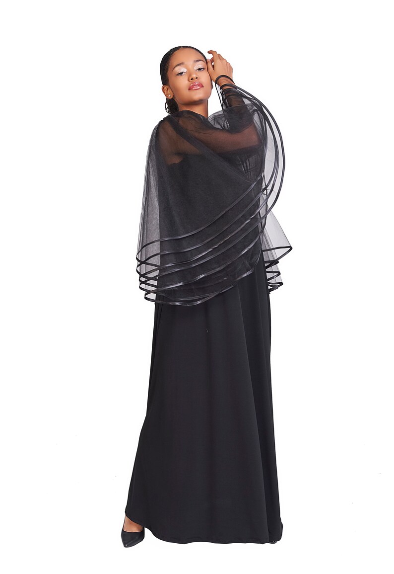 May include: A floor-length black dress featuring a sheer, layered tulle capelet overlay. The capelet is constructed from multiple tiers of black mesh fabric, each finished with a solid black ribbon trim along the curved edges, creating a voluminous silhouette.