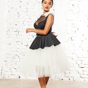 May include: A black sleeveless top with a sheer mesh neckline and white ruffled shoulder accents, paired with a black peplum waist and a voluminous white tulle midi skirt. The ensemble is completed with black high-heeled pumps.