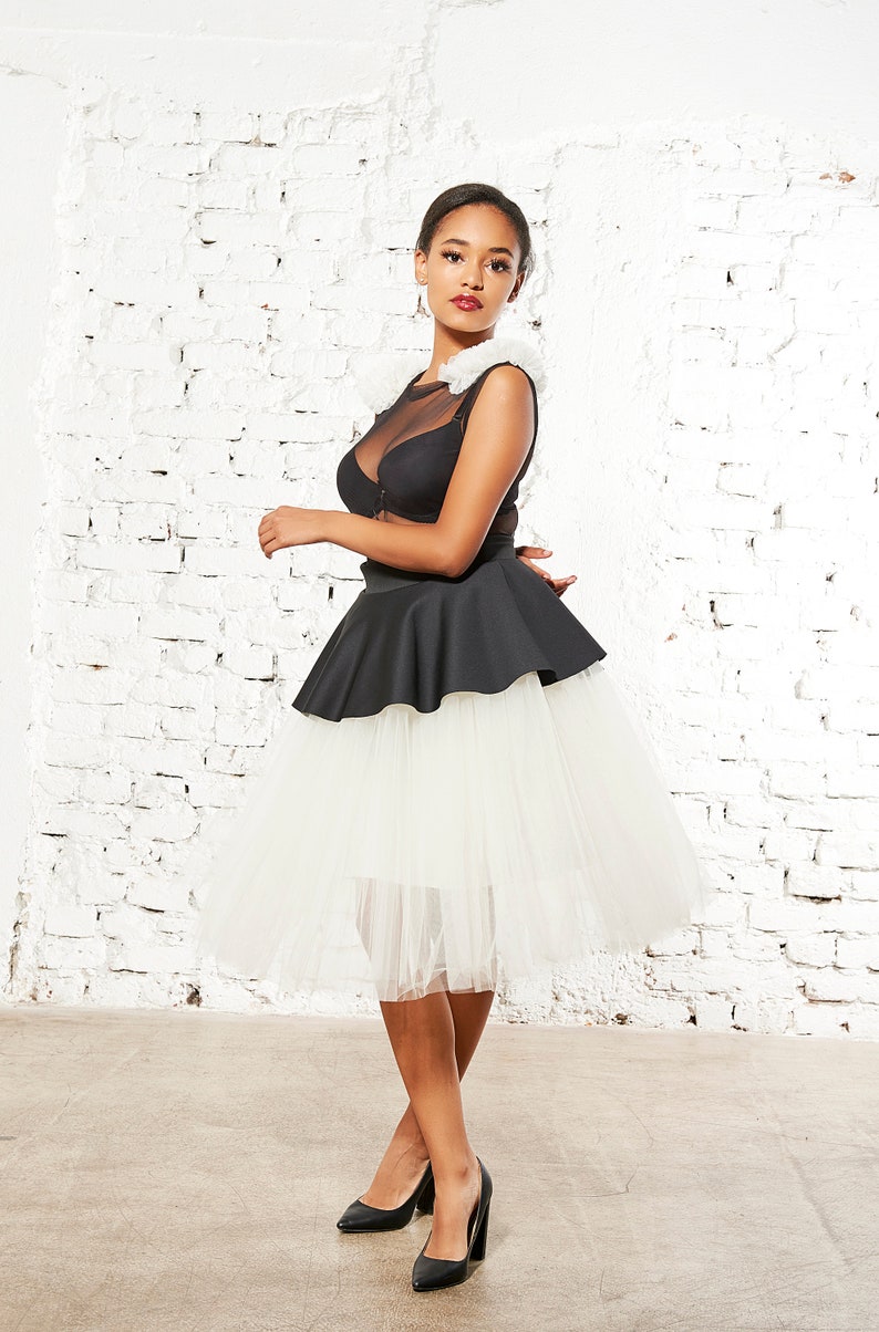 May include: A black sleeveless top with a sheer mesh neckline and white ruffled shoulder accents, paired with a black peplum waist and a voluminous white tulle midi skirt. The ensemble is completed with black high-heeled pumps.