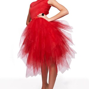 May include: A vibrant red dress featuring a sleeveless bodice and a voluminous, multi-layered tulle skirt that falls to a mid-calf length. The skirt has an asymmetrical, jagged hemline with sheer, overlapping fabric panels creating a textured effect.