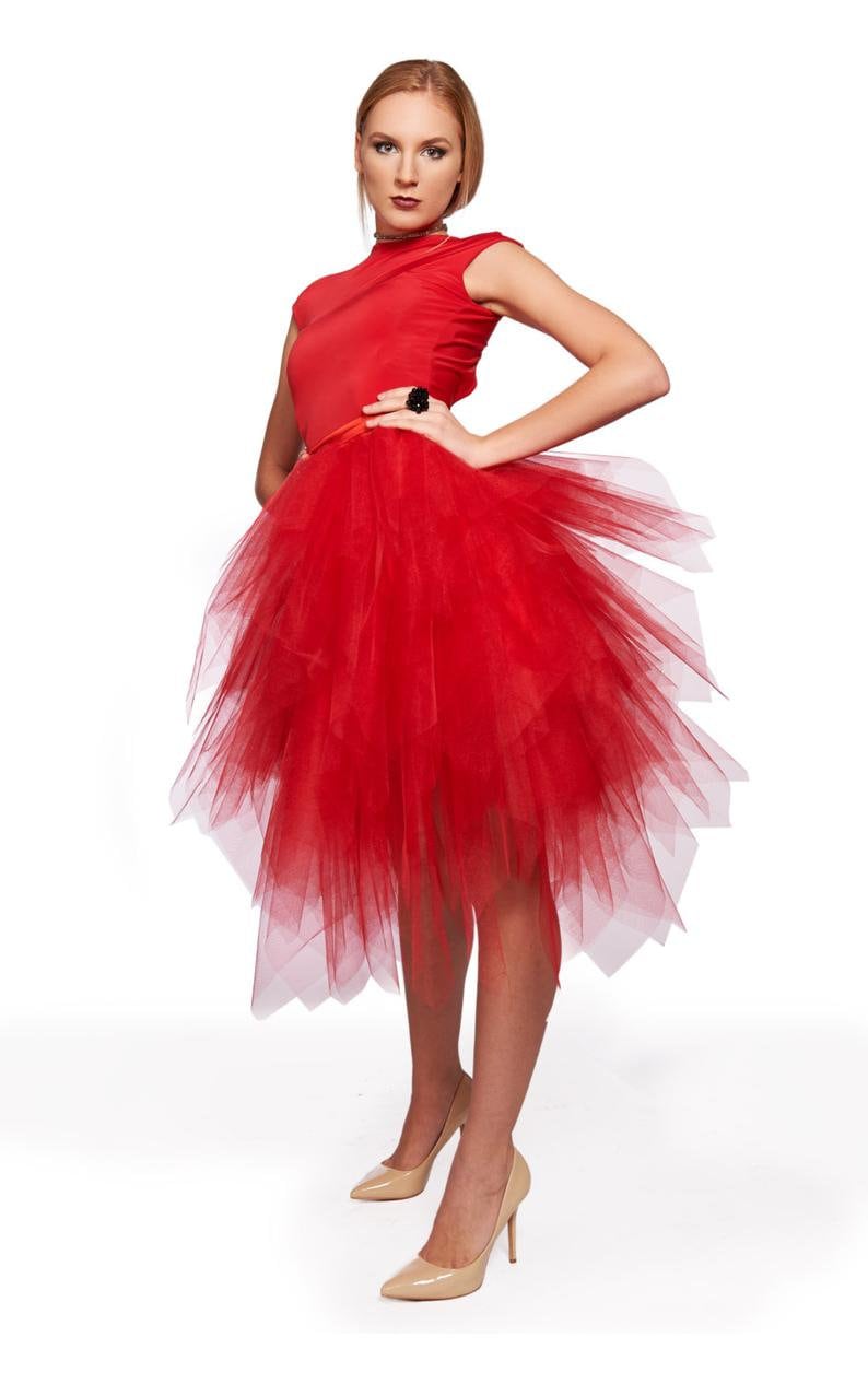 May include: A vibrant red dress featuring a sleeveless bodice and a voluminous, multi-layered tulle skirt that falls to a mid-calf length. The skirt has an asymmetrical, jagged hemline with sheer, overlapping fabric panels creating a textured effect.
