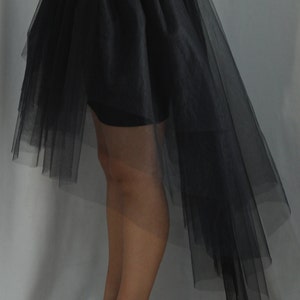 May include: A black high-low skirt featuring a wide, solid waistband and multiple layers of sheer, lightweight tulle fabric that cascades down to a longer length in the back, paired with red high-heeled pumps with a platform sole.