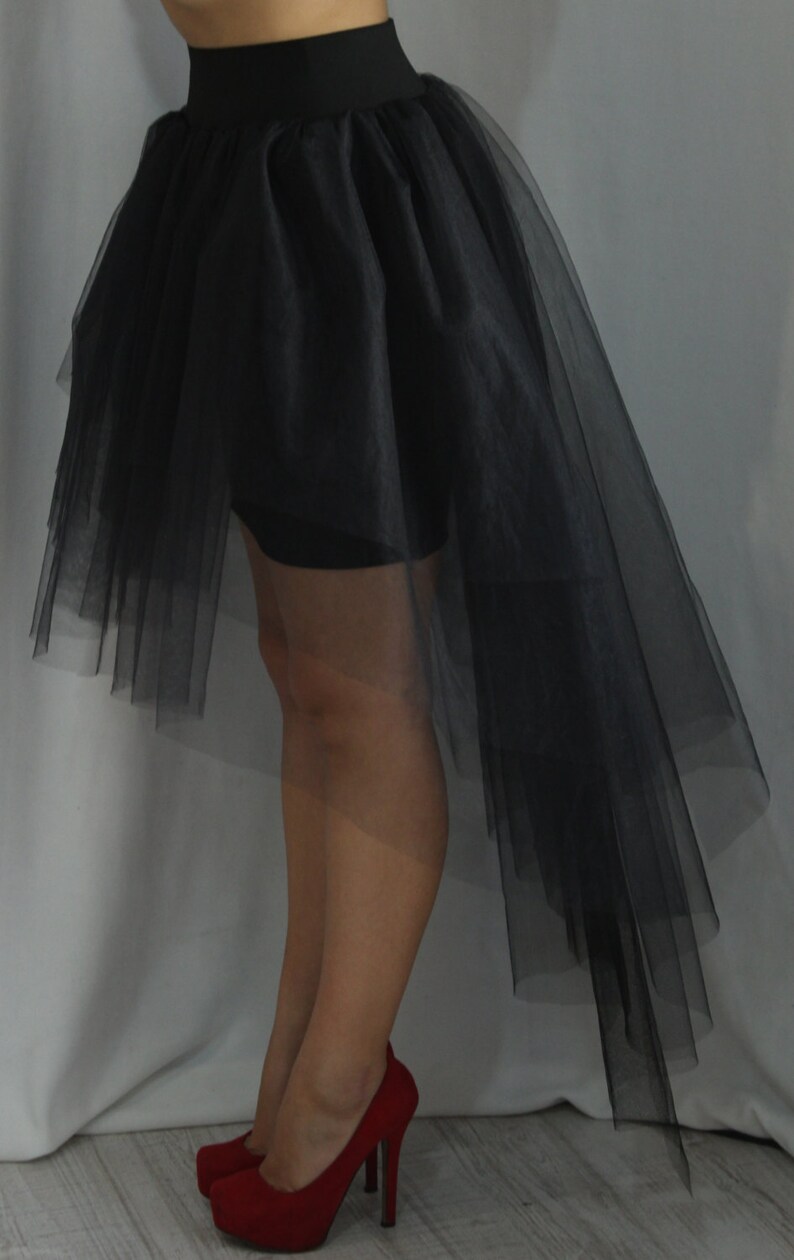 May include: A black high-low skirt featuring a wide, solid waistband and multiple layers of sheer, lightweight tulle fabric that cascades down to a longer length in the back, paired with red high-heeled pumps with a platform sole.