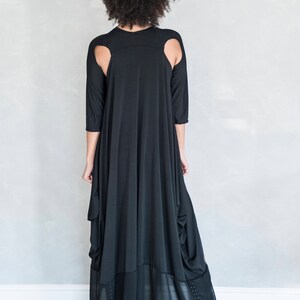 May include: A black floor-length dress featuring three-quarter length sleeves, shoulder cutouts, and a draped silhouette. The lower portion of the garment includes a sheer panel with subtle vertical detailing along the sides of the hemline.